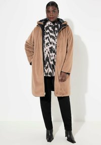 Beige nepbont lange jas met een zwarte ritssluiting en capuchon, gedragen over een gebreide trui, gecombineerd met zwarte broek en enkellaarsjes.