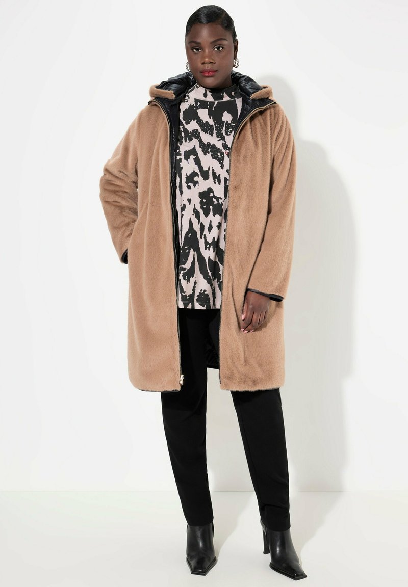 Beige nepbont lange jas met een zwarte ritssluiting en capuchon, gedragen over een gebreide trui, gecombineerd met zwarte broek en enkellaarsjes.