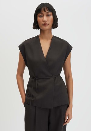 Vrouw met kort donker haar die een mouwloos zwart wikkeltopje draagt met een dunne riem, staand met één hand in de zak tegen een witte achtergrond.