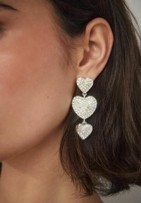 Das Ohr einer Frau, die einen hängenden Ohrring trägt, der aus drei funkelnden silbernen Herzen in unterschiedlicher Größe besteht, die vertikal miteinander verbunden sind.