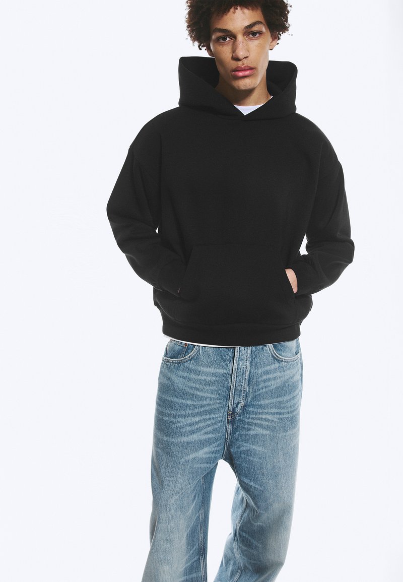 Jeune personne portant un sweat à capuche noir, mains dans la poche avant, et un jean ample bleu clair, debout devant un fond blanc.