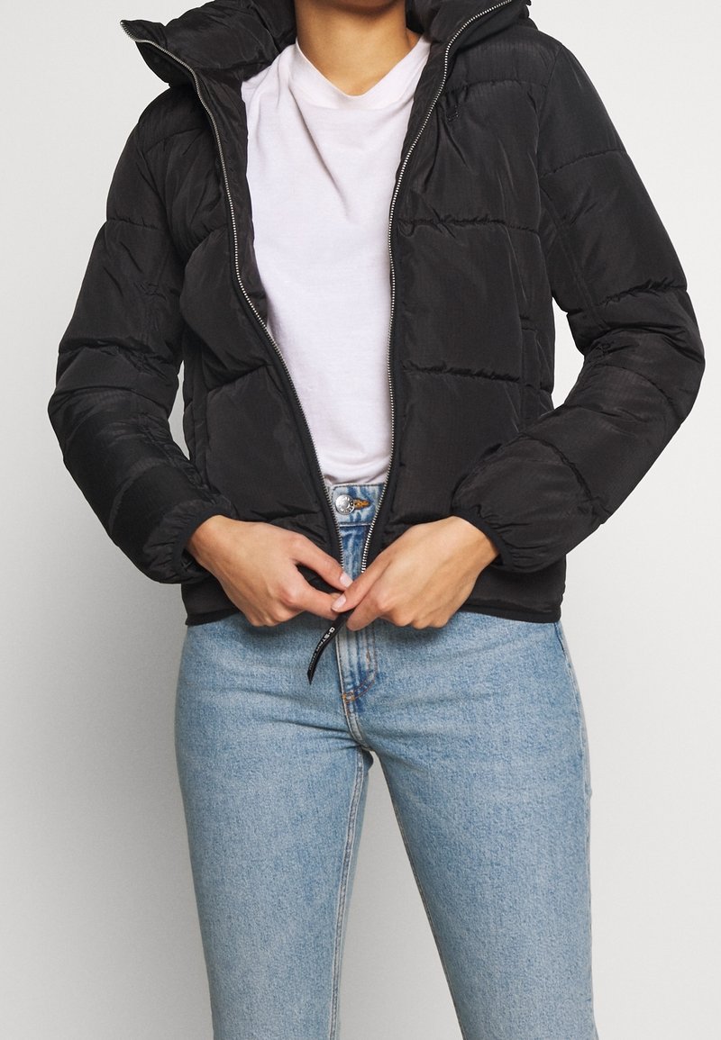 Doudoune noire avec fermeture éclair devant, capuche et poignets élastiques. Texture légèrement matelassée. Le mannequin porte un t-shirt blanc et un jean bleu.