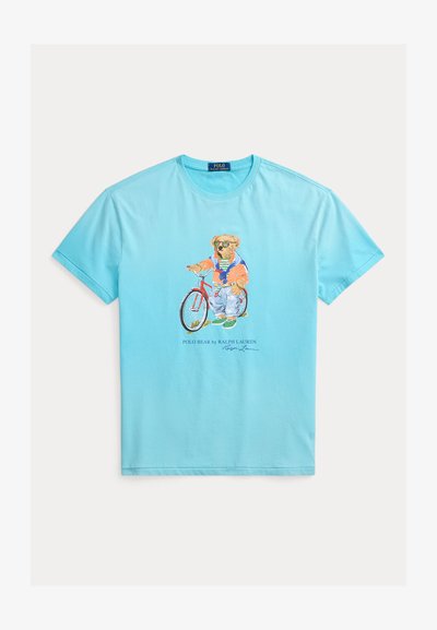 Ανοιχτό μπλε μπλουζάκι με γραφικό ενός αρκούδου που φορά γυαλιά ηλίου και εσώρουχα καθημερινής χρήσης, στέκεται δίπλα σε ένα κόκκινο ποδήλατο.