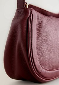 Gros plan d'un sac à main en cuir rouge foncé texturé avec un rabat arrondi et des détails cousus.