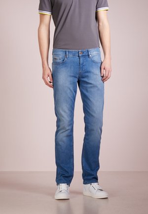 Mand iført falmede blå jeans, hvide sneakers og en grå kortærmet skjorte med gul og hvid ærmekant, står mod en ensfarvet baggrund.
