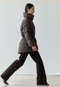Kurtka puchowa w ciemnobrązowym kolorze z wysokim kołnierzem, taliowana, z kontrastową fakturą. Połączona z brązowymi spodniami o szerokich nogawkach i czarnymi butami.