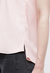 Gros plan sur le côté d'une personne portant un chemisier rose clair avec une fente sur le côté et un jean gris foncé.