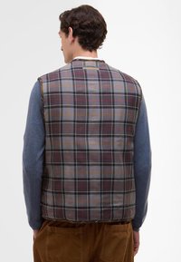 Kostkovaná polstrovaná vesta s kombinací šedých, červených a oranžových tónů, pod ní modrá košile s dlouhým rukávem. Tkanina má hladkou strukturu.