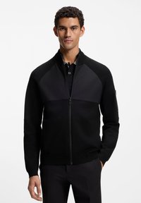 Μαύρο μπουφάν με φερμουάρ, με υφασμάτινη επάνω επιφάνεια, λεία κάτω ενότητα, όρθιο γιακά και διακριτικές λεπτομέρειες μεταλλικών στοιχείων. Φορεμένο πάνω από μαύρο πόλο.