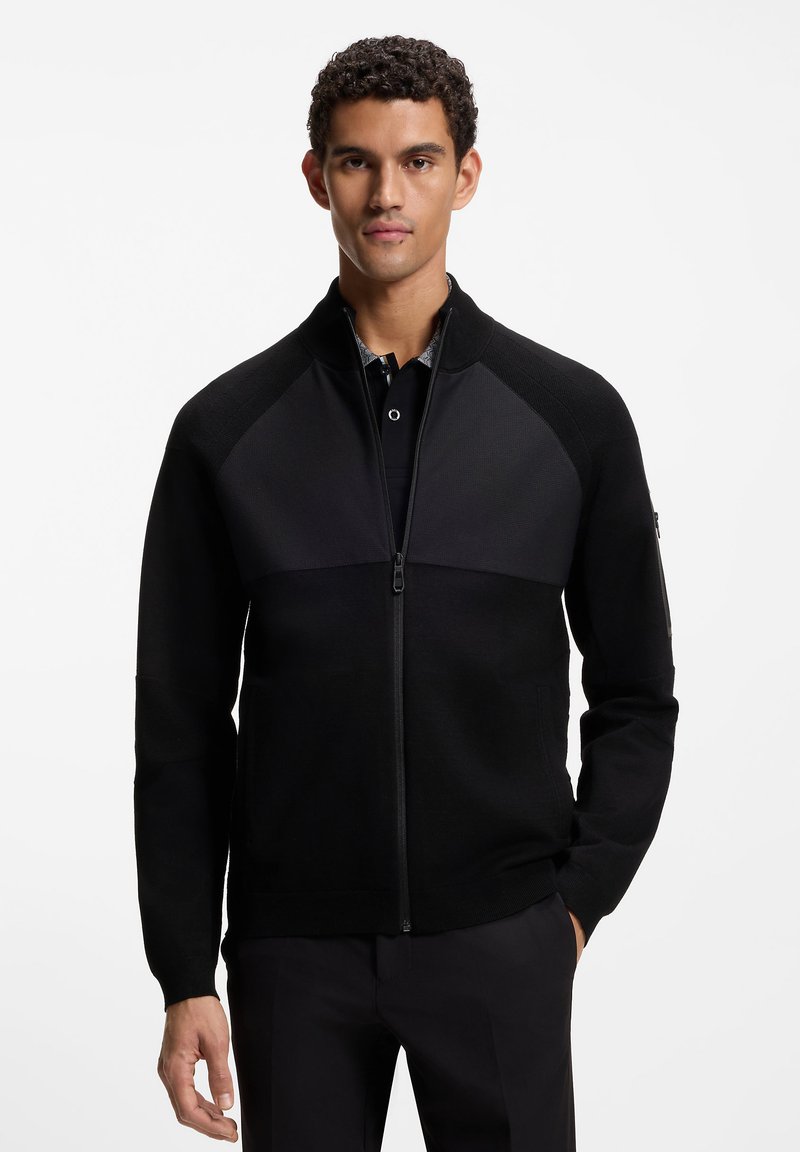 Μαύρο μπουφάν με φερμουάρ, με υφασμάτινη επάνω επιφάνεια, λεία κάτω ενότητα, όρθιο γιακά και διακριτικές λεπτομέρειες μεταλλικών στοιχείων. Φορεμένο πάνω από μαύρο πόλο.