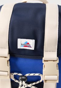 Σακίδιο από ύφασμα σε ναυτικό και μπλε χρώμα με μπεζ ιμάντες, λευκή ετικέτα με πολύχρωμο λογότυπο και κορδόνι σύσφιξης στο μπροστινό μέρος.