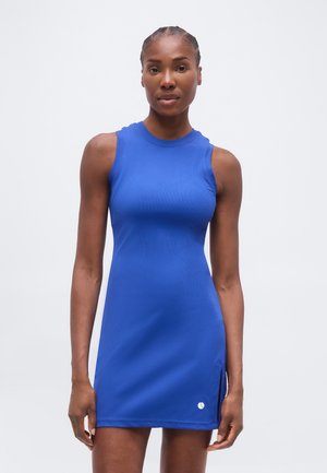 Γυναίκα που φοράει αμάνικο μπλε αθλητικό φόρεμα με μικρό λογότυπο κοντά στο τελείωμα, στεκόμενη μπροστά σε ένα απλό φωτεινό φόντο.