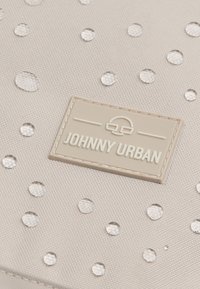 Világos bézs táska, texturált felülettel, körönként elhelyezett átlátszó díszítésekkel és egy téglalap alakú logóval, amelyen az áll: "JOHNNY URBAN."