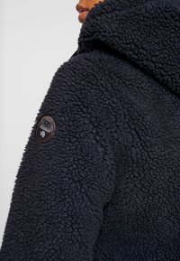 Marineblaue Fleecejacke mit strukturiertem Außenmaterial, einer Kapuze und einem runden Logopatch auf der Schulter. Weiches und warmes Material.