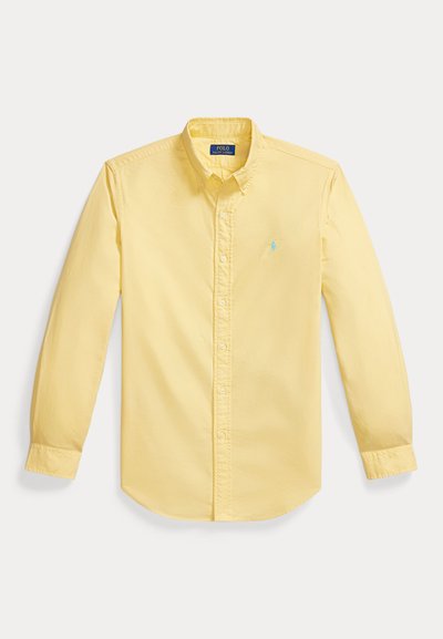 Chemise jaune à manches longues avec boutons et col, avec un logo brodé bleu clair sur la poitrine gauche, posée à plat sur un fond blanc.