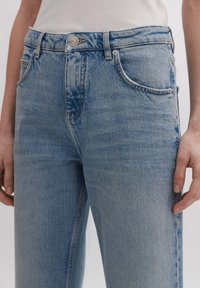 Jeans de mezclilla azul claro con cintura alta, cinco bolsillos, botón plateado y detalles de costura. Textura suave con un acabado ligeramente descolorido.
