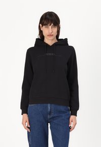 Felpa con cappuccio nera realizzata in tessuto morbido, con logo frontale, cordini e polsini a coste. Abbinata a jeans di denim blu.