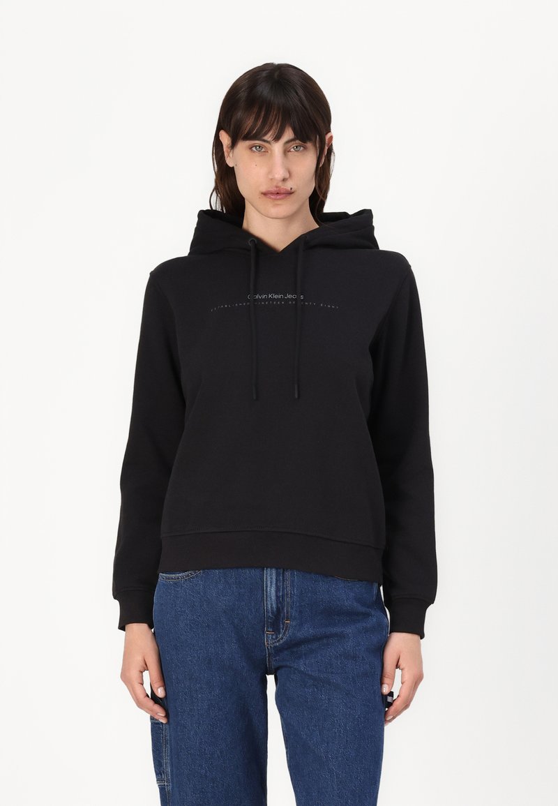 Felpa con cappuccio nera realizzata in tessuto morbido, con logo frontale, cordini e polsini a coste. Abbinata a jeans di denim blu.