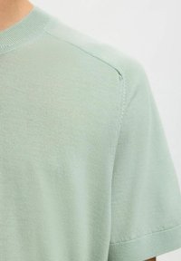 Gros plan d'un pull tricoté vert clair à manches courtes montrant la couture de l'épaule ainsi que le col, les poignets des manches côtelés.