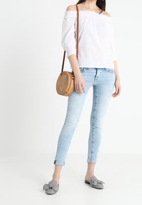 Top bianco a spalle scoperte, jeans skinny azzurri chiari, mocassini grigi a fantasia e una borsa rotonda intrecciata con una tracolla marrone.