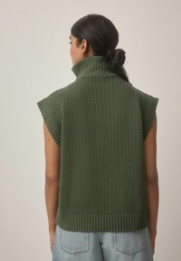 Ärmelloser Strickpullover in Dunkelgrün mit hohem Kragen, ausgestattet mit einem strukturierten Muster und geripptem Saum, kombiniert mit hellblauen Jeans.