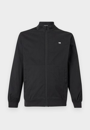 Chaqueta negra con cremallera, cuello alto, puños elásticos, bolsillos laterales y una pequeña placa rectangular con logo en el pecho.