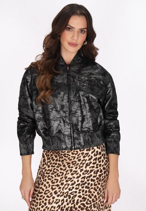 Vrouw met lang golvend haar, gekleed in een glanzend zwart jasje met rits en een rok met luipaardprint, staand tegen een witte achtergrond.