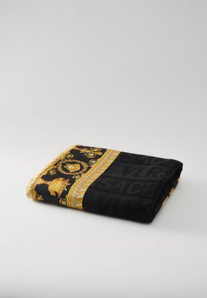 Melns pleds ar zelta ornamentālu dizainu, ietverot baroka rakstu un vārdu "VERSACE", kas integrēts gar malu. Mīksta tekstūra.
