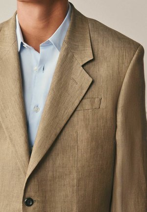 Man wearing light brown textured blazer over a light blue dress shirt with neck and upper chest visible.