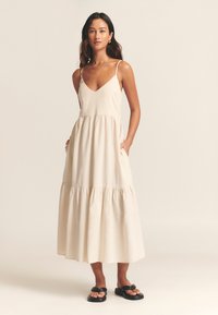 Light beige maxi dress with a v-neck and spaghetti straps, featuring a tiered skirt and pockets. Model wears black sandals.