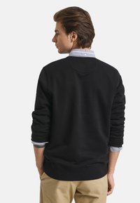 Black sweatshirt with crew neck, ribbed cuffs, and waistband, layered over a light blue collared shirt, worn with khaki pants.