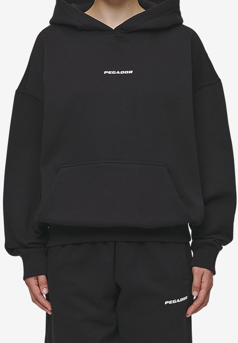 Personne portant un sweat à capuche noir oversize et un pantalon noir assorti, tous deux avec le logo blanc "PEGADOR" au centre avant et sur la cuisse gauche.