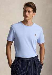 Jeune homme portant un T-shirt bleu clair à manches courtes et un pantalon bleu marine à fines rayures, debout les mains dans les poches, regardant de côté.