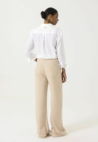 Camisa blanca de botones con cuello y puños enrollados, combinada con unos pantalones anchos beige que cuentan con bolsillos traseros y una textura sutil.