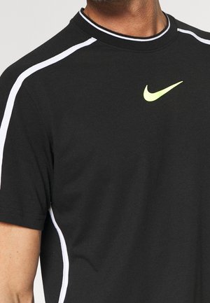 Detailní záběr na osobu, která má na sobě černé tričko Nike s bílými pruhy na ramenou a neonově žlutým logem Nike swoosh na hrudi.