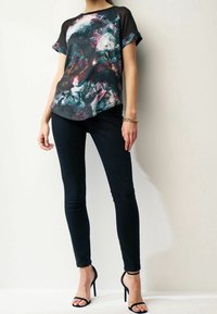 Vícebarevná blůza s abstraktním vzorem a krátkými rukávy, se zaobleným lemem, doplněná o přiléhavé černé džíny a černé sandály na podpatku.