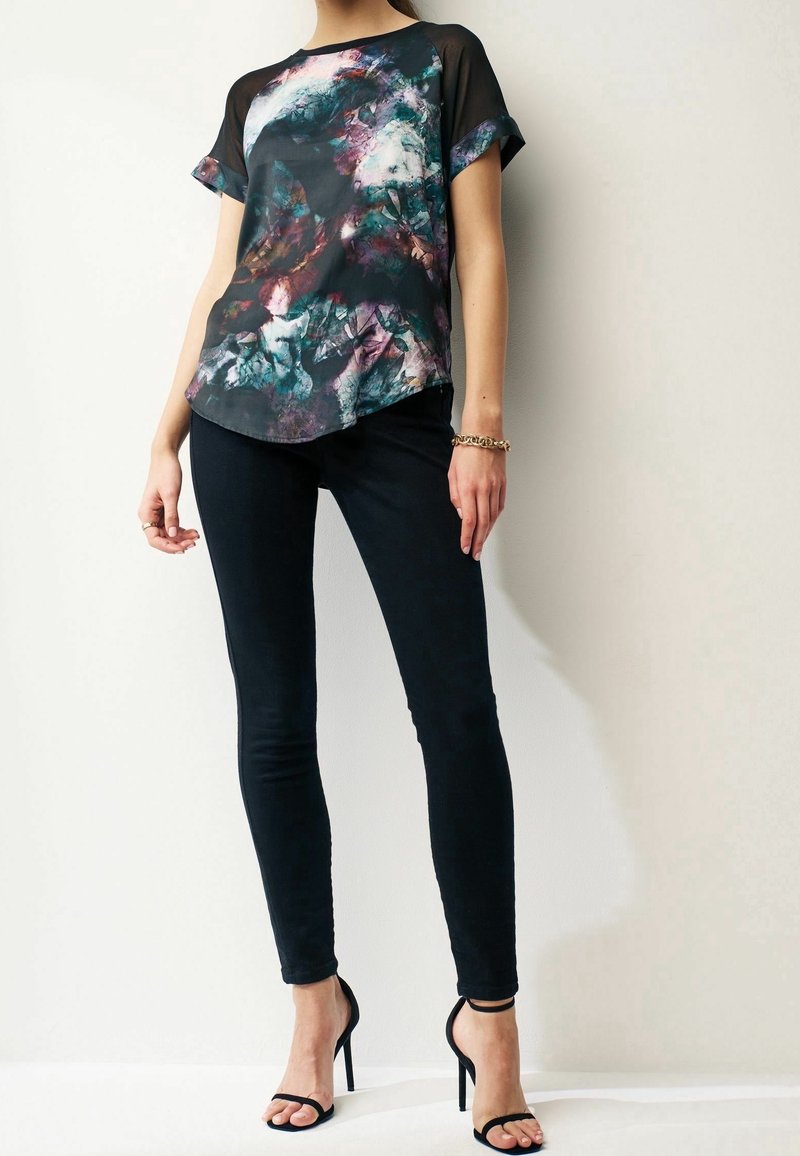 Vícebarevná blůza s abstraktním vzorem a krátkými rukávy, se zaobleným lemem, doplněná o přiléhavé černé džíny a černé sandály na podpatku.