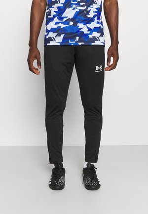 Mężczyzna w czarnych spodniach sportowych Under Armour, czarno-białych butach sportowych i niebieskiej koszuli w biały wzór, stoi na białym tle.