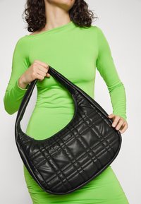 Sac hobo matelassé noir avec une anse courbée, en cuir brillant. Le sac présente une texture lisse et une fermeture éclair pour la fonctionnalité.