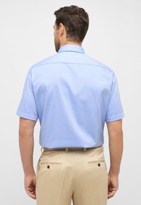 Een lichtblauw, kortmouwig overhemd met een klassieke kraag, een gladde textuur en opgerolde mouwen, gecombineerd met beige broeken met achterzakken.