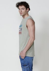 Bez piedurknēm, gaiši zaļa kokvilnas tvertnes krekls ar grafisku attēlu zilos un sarkanos toņos. Tam ir brīvs piegriezums un noapaļots apakšmalas dizains, kas saskaņots ar ziliem cargo biksēm.