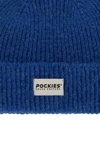Modrá pletená čepice s žebrovanou texturou, se bílou nášivkou s nápisem "POCKIES COUCH COUTURE". Tlustý, teplý materiál.