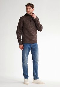 Hnědý pletený svetr s vysokým límcem a žebrovanými manžetami, v kombinaci s modrými džíny a bílými teniskami, na jednobarevném pozadí.