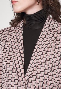 Blazer s geometrickým vzorem v růžových, černých a šedých tónech. Strukturovaný design s klopami, vrstvený přes černý rolák. Měkká textura.