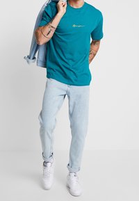 Tyrkysové bavlněné tričko s kulatým výstřihem, zdobené žlutě vyšitým logem, v kombinaci se světle modrými džíny a bílými teniskami.