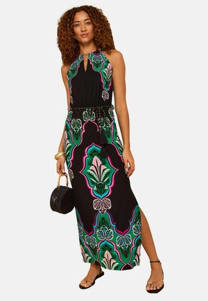 Naisel kiharate juuste ja musta varrukateta maksikleit, millel on rohelised, roosad ja sinised lillemustrid, hoiab ümmargust musta käekotti ja kannab sandaale.