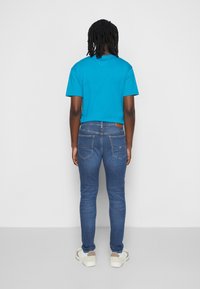 Camiseta turquesa de manga corta combinada con jeans azul oscuro ajustados. Bolsillos traseros visibles y una parche de cuero marrón en la cintura de los jeans.