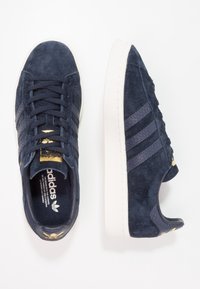 Námořnicky modré semišové tenisky s třemi texturovanými pruhy. Bílá gumová podrážka a zlaté logo. Kulatá špička a design na zavazování tkaniček. Plochý podpatek.