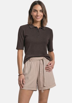 Femme portant un polo à manches courtes côtelé marron foncé et un short beige clair ceinturé, debout devant un fond uni.