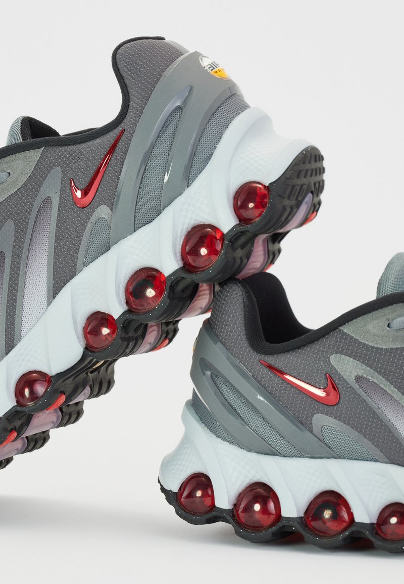 Zapatos deportivos grises con una parte superior de malla, acentos rojos y burbujas de amortiguación distintivas en una suela blanca, con un diseño curvado.